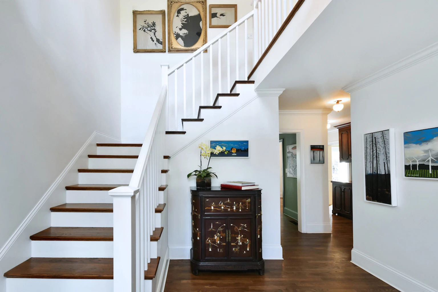 Listing photo of a bright entry hall with a wooden staircase, framed artwork and an inlaid cabinet